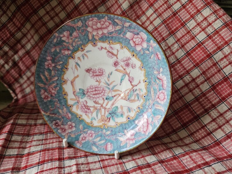 Op de afbeelding: Een vintage porseleinen schotel met een blauw en wit bloemenmotief. De schotel heeft roze bloemen en groene bladeren op een lichtblauwe achtergrond. De rand van de schotel is versierd met een gouden rand.