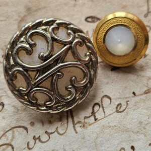 May include: Two vintage buttons with a white pearl-like center. The button on the left has a silver filigree design and a rope-like border. The button on the right has a gold border and a smooth surface.