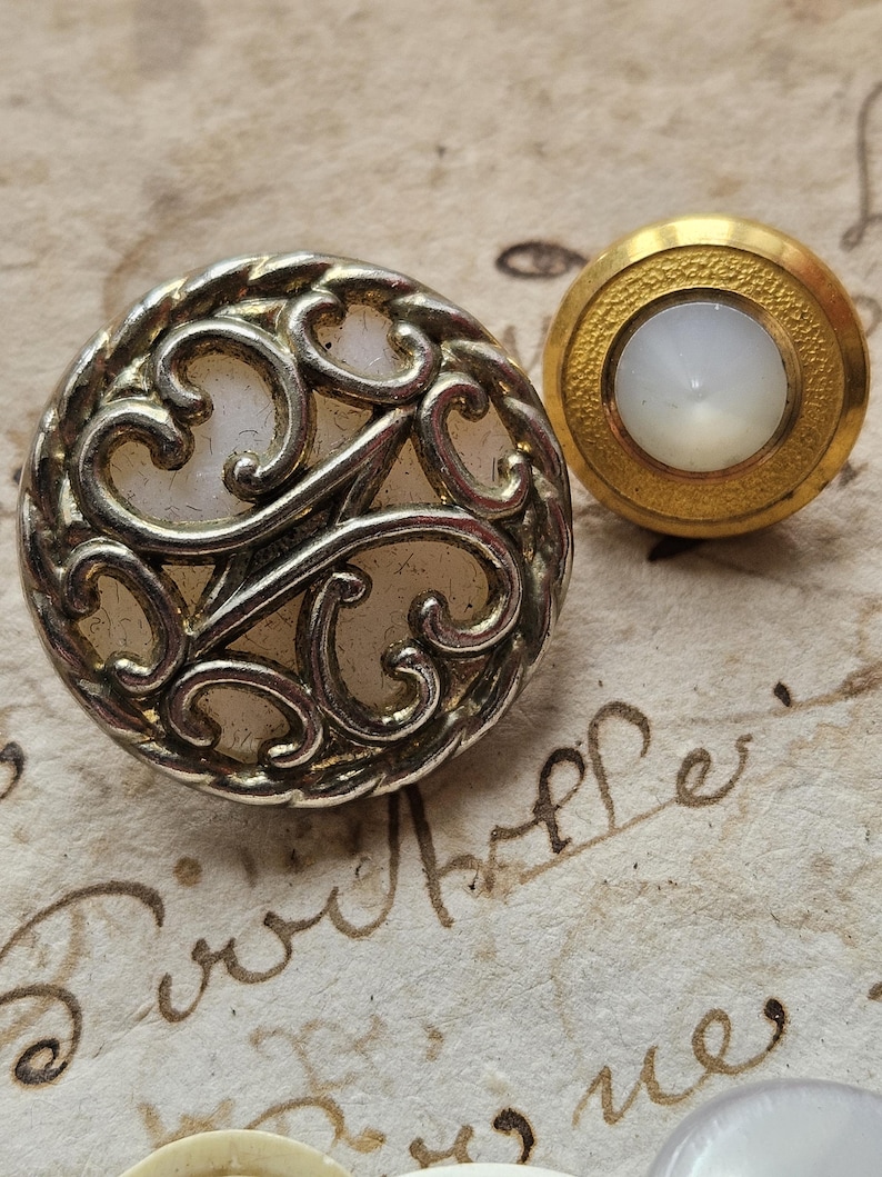 May include: Two vintage buttons with a white pearl-like center. The button on the left has a silver filigree design and a rope-like border. The button on the right has a gold border and a smooth surface.