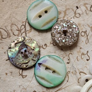 May include: Four vintage buttons with different designs and colors. Two buttons are white with green stripes and two holes. One button is iridescent with a brown background and a floral design. The last button is brown with iridescent glitter.