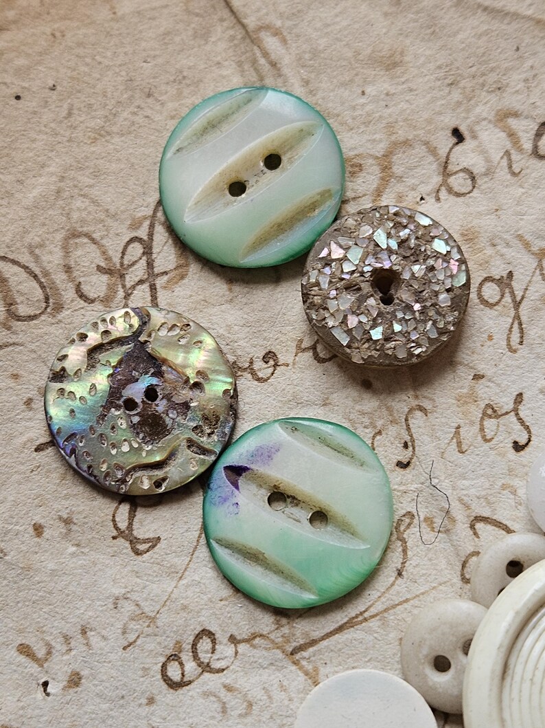 May include: Four vintage buttons with different designs and colors. Two buttons are white with green stripes and two holes. One button is iridescent with a brown background and a floral design. The last button is brown with iridescent glitter.