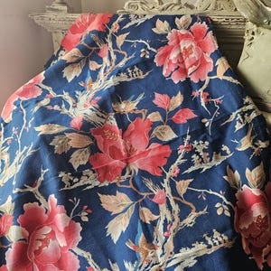 Op de afbeelding: Een marineblauwe stof met een bloemenpatroon met grote rode pioenen, kleinere roze bloemen en vogels. Het ontwerp bevat takken en bladeren in tinten beige en bruin. De stof is gedrapeerd over een witte, sierlijke plank.