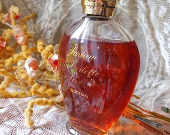 Può includere: Un flacone di profumo vintage in vetro con un tappo dorato. Il flacone è riempito con un liquido ambrato scuro e ha il testo "Parfum de Colette Paris ROCHAS" sull'etichetta.