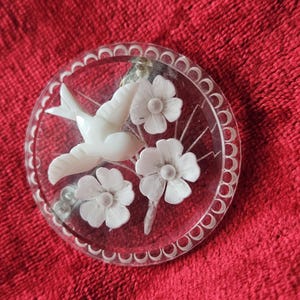 May include: A clear, round brooch with a white bird and three white flowers. The bird is in flight, and the flowers are intricately detailed. The brooch has a scalloped edge and is set against a red background.