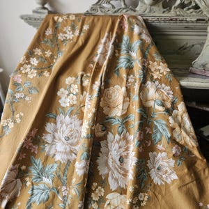 May include: A mustard yellow fabric featuring a repeating floral pattern with large white and cream peonies, smaller light pink blossoms, and muted sage green foliage. The textile is draped, showing soft folds and a matte finish throughout.