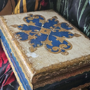 May include: An antique, rectangular box with a cream-colored lid featuring a blue and gold floral design. The box has gold trim and blue accents on the sides. The box is placed on a colorful patterned fabric.