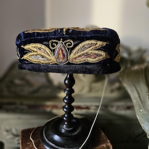 May include: A dark blue velvet hat with gold and pink embroidery. The hat is on a wooden stand.