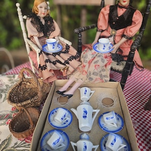 May include: A miniature porcelain tea set in a cardboard box, featuring a white teapot, creamer, and four cups with saucers, all decorated with blue sailboat motifs. Two dolls in vintage-style clothing sit nearby with two small woven wicker baskets.