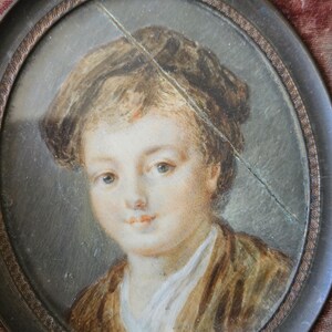 May include: Oval portrait miniature of a young person with light skin, wearing a brown jacket and white shirt. The portrait is set in a dark, ornate frame. The painting has a crack across the surface.