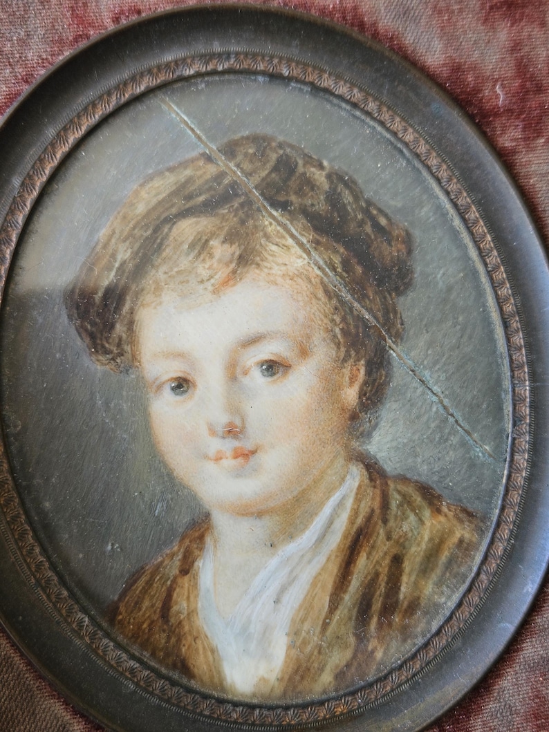 May include: Oval portrait miniature of a young person with light skin, wearing a brown jacket and white shirt. The portrait is set in a dark, ornate frame. The painting has a crack across the surface.