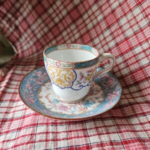 Op de afbeelding: Een vintage theekopje en schotelset met een bloemenpatroon in tinten blauw, roze, geel en groen. Het kopje en de schotel hebben een gouden rand.