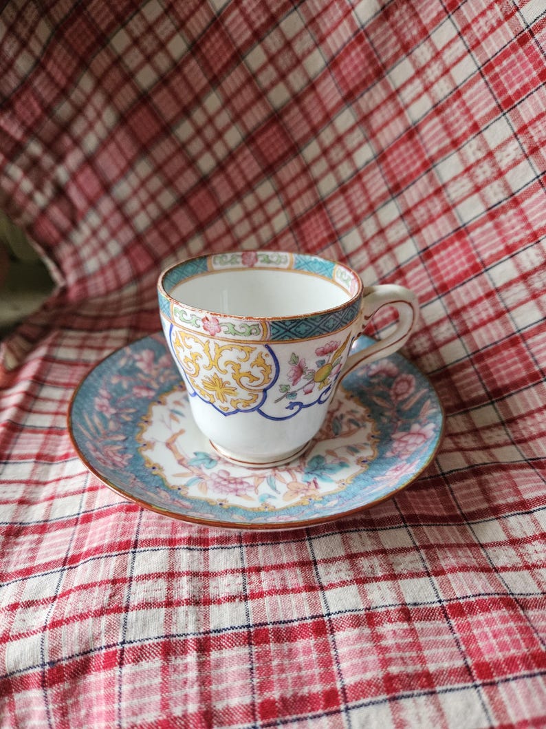 Op de afbeelding: Een vintage theekopje en schotelset met een bloemenpatroon in tinten blauw, roze, geel en groen. Het kopje en de schotel hebben een gouden rand.