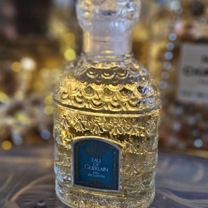 May include: A clear glass perfume bottle with a textured bee and scale pattern on the surface. The bottle features a rounded stopper and a blue rectangular label with the text EAU DE GUERLAIN eau de toilette printed in white and gold lettering.