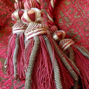 May include: Close-up of decorative tassels in shades of red, gold, and cream. The tassels feature intricate detailing with twisted cords, metallic accents, and decorative ball-shaped elements. The background is a patterned red fabric.