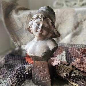Puede incluir: Un busto vintage de un niño con un sombrero. El busto está hecho de yeso y pintado en un color marrón claro. La base del busto está inscrita con las palabras "Sur La Falaise".