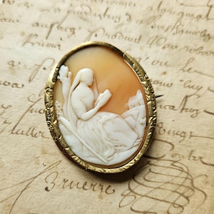 May include: Oval cameo brooch with a gold-tone frame. The cameo features a carved relief of a woman reading, set against a peach-colored background. The woman is wearing a draped gown. The brooch is pinned to a vintage paper background.
