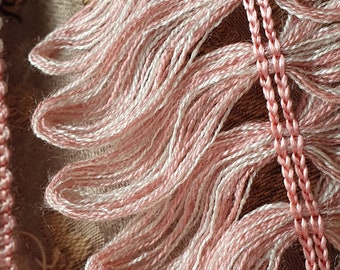 May include: Close-up of pink and white embroidery floss, with a braided edge detail. The floss is arranged in wavy bundles, creating a textured effect. The background is a muted, patterned fabric.