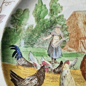 May include: A vintage ceramic plate with a hand-painted scene of a woman feeding chickens in a farmyard. The plate features a blue and white rooster, brown and white chickens, and a woman in a blue dress and white apron. The background includes a green field, trees, and a brown barn.