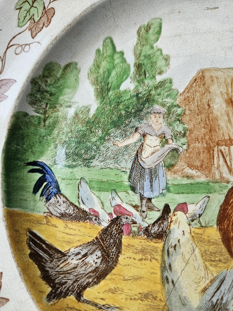 May include: A vintage ceramic plate with a hand-painted scene of a woman feeding chickens in a farmyard. The plate features a blue and white rooster, brown and white chickens, and a woman in a blue dress and white apron. The background includes a green field, trees, and a brown barn.