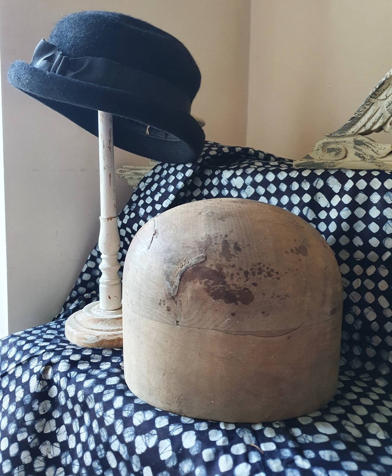 May include: A wooden hat block, also known as a hat form, is a tool used by milliners to shape hats. The block is made of wood and has a rounded top and a flat base. It is shown on a black and white patterned fabric.