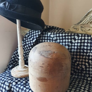 May include: A wooden hat block, also known as a hat form, is a tool used by milliners to shape hats. The block is made of wood and has a smooth, rounded surface. It is sitting on a black and white patterned fabric.
