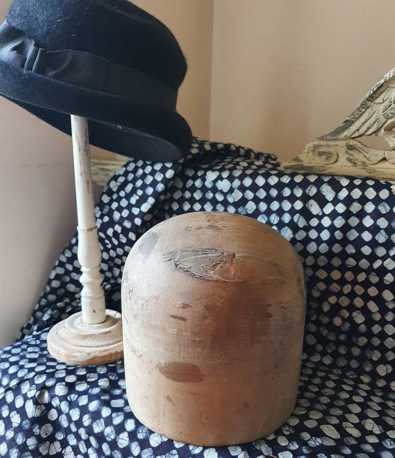 May include: A wooden hat block, also known as a hat form, is a tool used by milliners to shape hats. The block is made of wood and has a smooth, rounded surface. It is sitting on a black and white patterned fabric.