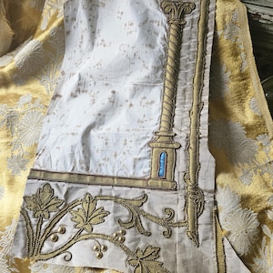 Può includere: Tessuto color crema con ricami dorati raffiguranti una colonna, foglie e viti. Il tessuto presenta un motivo maculato ed è posto su uno sfondo floreale dorato. Il ricamo è dettagliato e ornato, con un design classico.