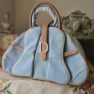 May include: Light blue denim handbag with tan leather accents. Features a silver metal "D" logo and tan leather handles. The bag has a rounded shape and is set on a floral patterned surface.