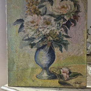 May include: Oil painting of a floral still life. A blue vase holds a bouquet of white and pink flowers with green leaves. A single flower and leaves lie on the table. The painting has a textured appearance and is signed "J. Trebaul".