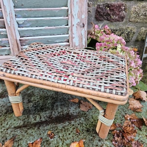 Op de afbeelding: Een kleine, geweven, houten kruk met een lichtblauwe en rode geweven bovenkant. De kruk staat op een bruin oppervlak met gevallen bladeren.