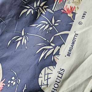 May include: Close-up of a fabric with a blue background and white bamboo and floral patterns. The fabric also features pink and red flowers. A white strip on the right side has the text "BERGAMOTTE" and "HOULES" printed on it.