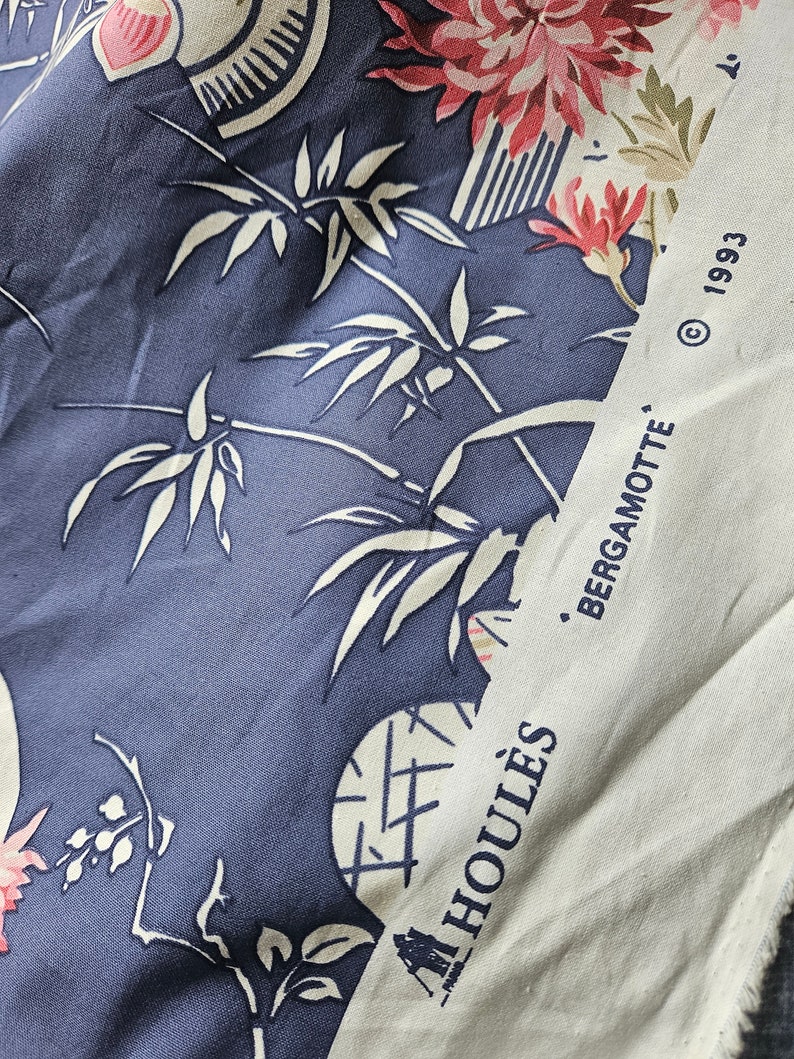 May include: Close-up of a fabric with a blue background and white bamboo and floral patterns. The fabric also features pink and red flowers. A white strip on the right side has the text "BERGAMOTTE" and "HOULES" printed on it.