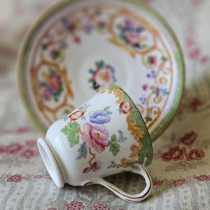 Op de afbeelding: Een witte porseleinen theekop en bijpassende schotel met een kleurrijk bloemmotief in roze, blauw en oranje, geaccentueerd door ingewikkeld goudkleurig krulwerk en een zachtgroene rand langs de geschulpte randen van de schotel.