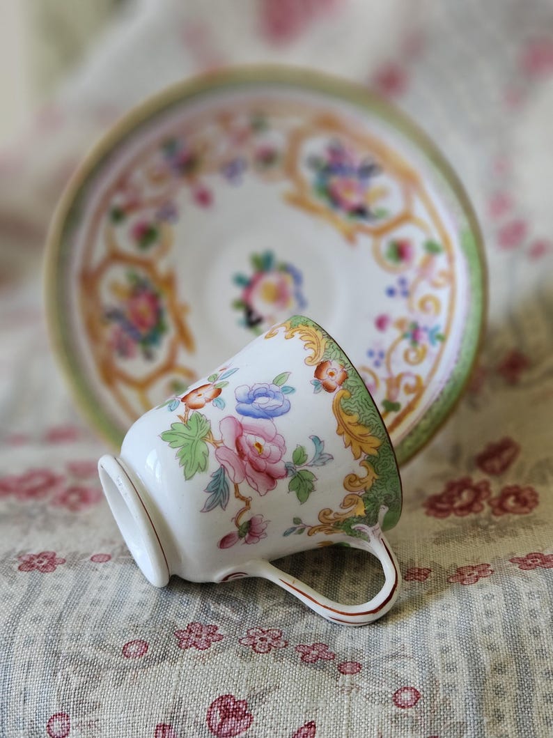 Op de afbeelding: Een witte porseleinen theekop en bijpassende schotel met een kleurrijk bloemmotief in roze, blauw en oranje, geaccentueerd door ingewikkeld goudkleurig krulwerk en een zachtgroene rand langs de geschulpte randen van de schotel.