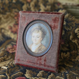 May include: A framed miniature portrait of a person with a light complexion, wearing a hat and a brown jacket. The portrait is set in an oval frame within a rectangular, burgundy velvet-covered case.