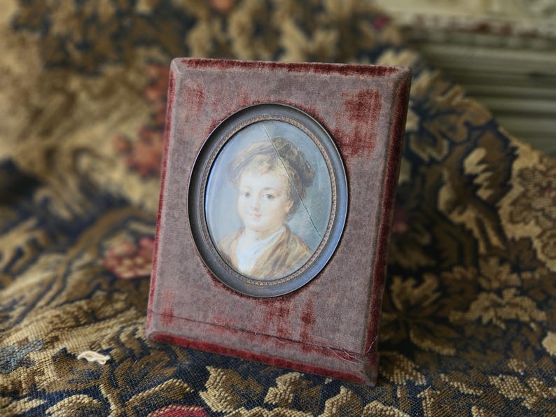 May include: A framed miniature portrait of a person with a light complexion, wearing a hat and a brown jacket. The portrait is set in an oval frame within a rectangular, burgundy velvet-covered case.