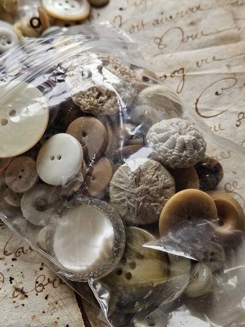 May include: A plastic bag filled with a variety of buttons in shades of white, brown, and beige. The buttons are a mix of different shapes and sizes, including round, square, and oval. Some of the buttons have a textured surface, while others are smooth.