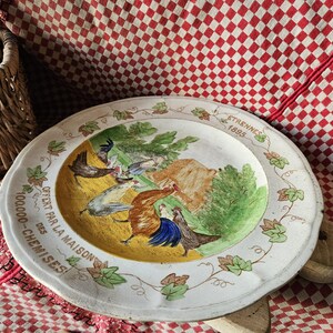 May include: A white ceramic plate with a yellow center and a green border. The plate features a painted scene of a rooster and hens in a farmyard. The text "OFFERT PAR LA MAISON 100,000 - CHEMISES!" is printed around the edge of the plate.