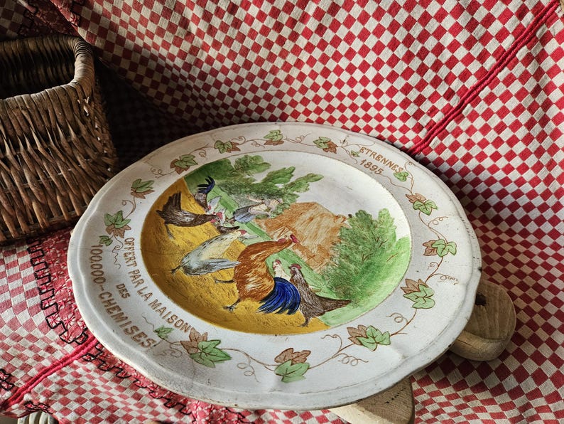 May include: A white ceramic plate with a yellow center and a green border. The plate features a painted scene of a rooster and hens in a farmyard. The text "OFFERT PAR LA MAISON 100,000 - CHEMISES!" is printed around the edge of the plate.