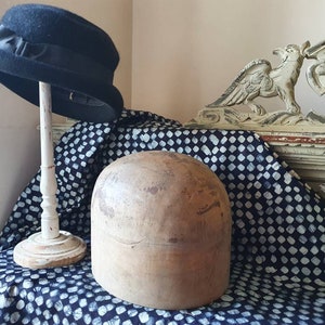May include: A wooden hat block, also known as a hat form, sits on a blue and white patterned fabric. A black felt hat with a black ribbon is on a white stand behind the hat block.