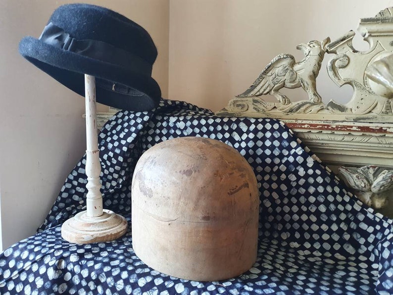 May include: A wooden hat block, also known as a hat form, sits on a blue and white patterned fabric. A black felt hat with a black ribbon is on a white stand behind the hat block.