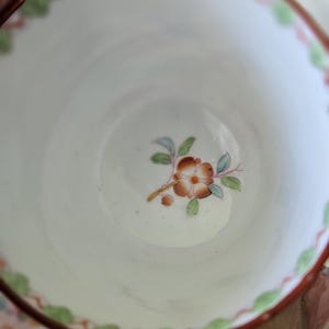 Op de afbeelding: Een witte keramische theekop met een handgeschilderd bloemmotief in het midden in bruine, groene en blauwe tinten. De rand is versierd met een geschulpte oranje rand en kleine groene bladaccenten, wat zorgt voor een gedetailleerd uiterlijk.