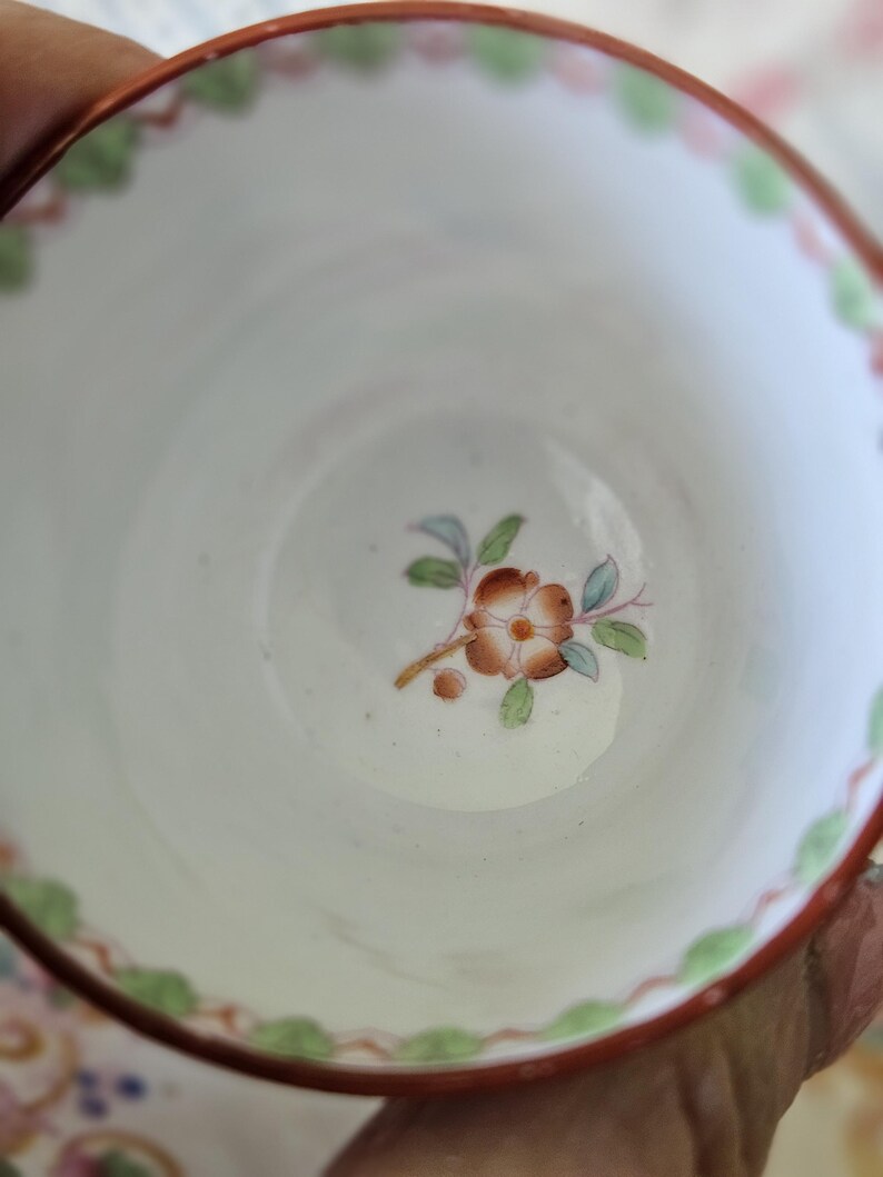 Op de afbeelding: Een witte keramische theekop met een handgeschilderd bloemmotief in het midden in bruine, groene en blauwe tinten. De rand is versierd met een geschulpte oranje rand en kleine groene bladaccenten, wat zorgt voor een gedetailleerd uiterlijk.