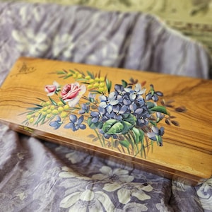 May include: A rectangular wooden box with a floral design. The box features a bouquet of blue hydrangeas, pink roses, and yellow mimosa. The box is made of light-colored wood with a visible wood grain.