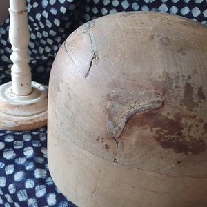 May include: A large, wooden hat form with a rounded top and a slightly flared base. The form is made of light brown wood and has several cracks and imperfections. The form is sitting on a blue and white patterned fabric.