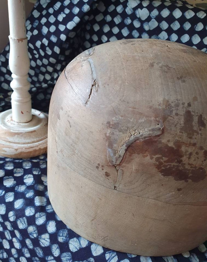 May include: A large, wooden hat form with a rounded top and a slightly flared base. The form is made of light brown wood and has several cracks and imperfections. The form is sitting on a blue and white patterned fabric.