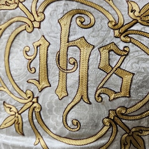 May include: Close-up of a cream-colored fabric with gold embroidery. The embroidery features the letters IHS in a decorative script, surrounded by ornate floral and scroll designs. The fabric has a subtle, textured pattern.