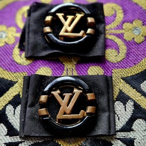 Può includere: Due accessori in tessuto nero, ciascuno con il logo "LV" dorato all'interno di una cornice circolare nera e dorata. Gli accessori sono su uno sfondo viola e oro.