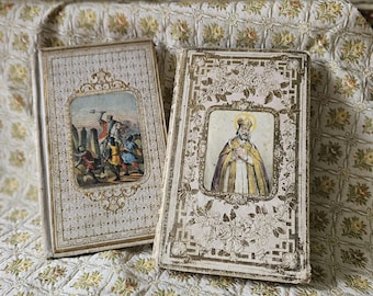 May include: Two antique, gold-trimmed, white books with ornate designs. One book features a religious image of a man in a robe and crown. The other book features a scene of people in medieval clothing.