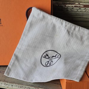 以下が含まれることがあります： 黒いロゴと茶色の紐が付いた、明るい灰色の巾着袋。 袋は「HERMES」の文字が印刷されたオレンジ色の箱の前に置かれています。 背景には他のオレンジ色の箱が含まれています。